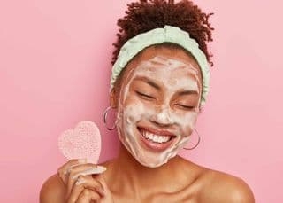 Beautiful optimistic Afro American woman cleanses face with foam, refreshes skin, has well cared complexion, holds heart shaped sponge for beauty procedures, stands bare shoulders with closed eyes